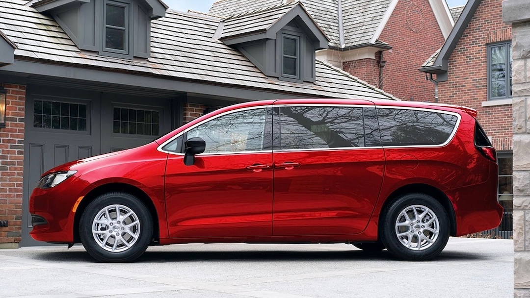 Profil latéral du Chrysler Grand Caravan 2025 rouge stationné devant une maison de banlieue, soulignant son espace familial.