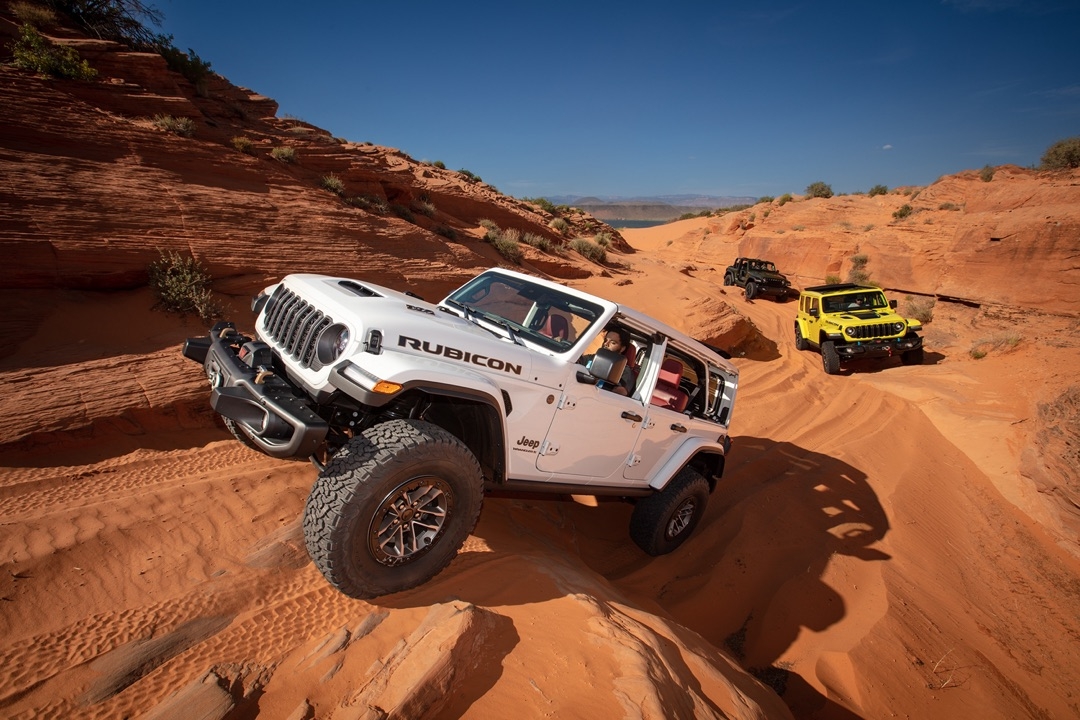 Jeep Wrangler Rubicon 2024 blanc escaladant une pente rocheuse dans le désert, accompagné d’un Rubicon jaune et d’un modèle noir en arrière-plan.