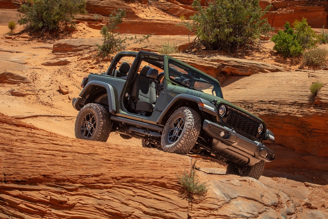 Jeep Wrangler Willys 2024 vert militaire franchissant un obstacle rocheux dans un désert, mettant en valeur sa garde au sol et ses pneus tout-terrain.