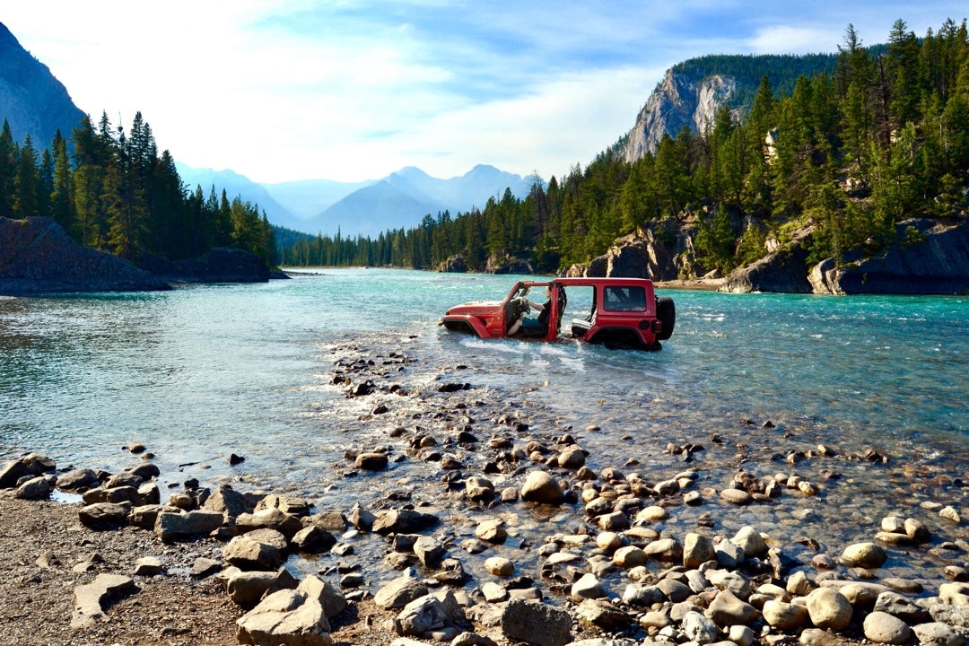 Jeep Wrangler Rubicon 2025 rouge traversant un gué peu profond, portes retirées, rochers au premier plan et montagnes boisées en arrière-plan.