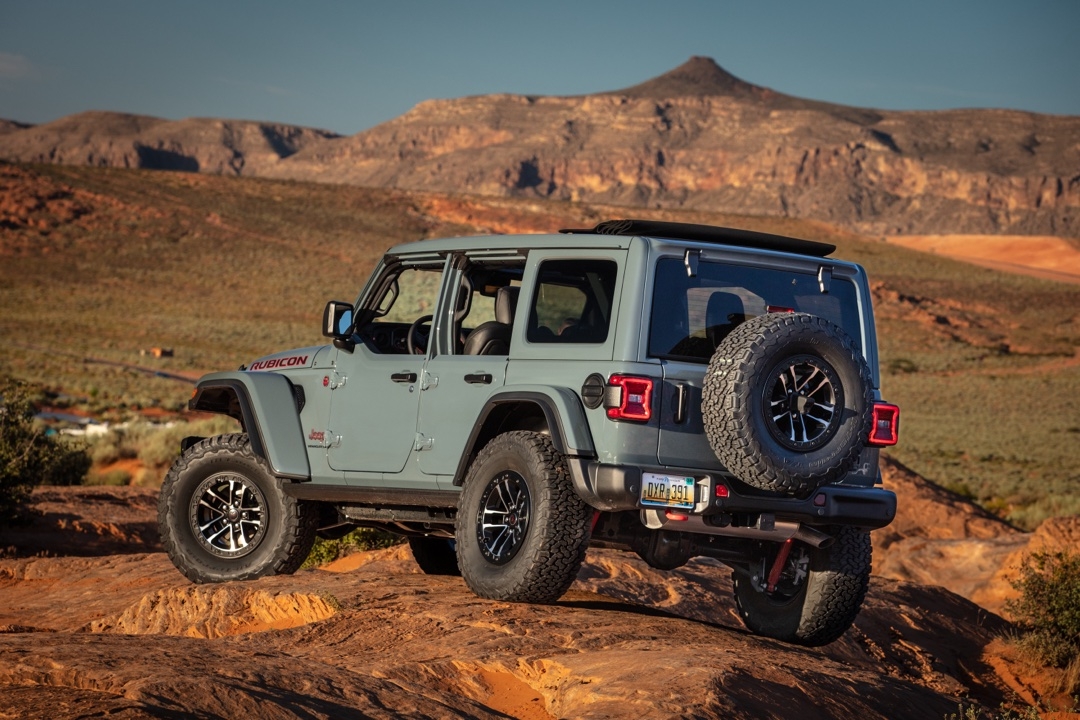 Vue arrière du Jeep Wrangler Rubicon 2025 gris sur terrain rocheux, avec roue de secours fixée et paysage montagneux à l’horizon.
