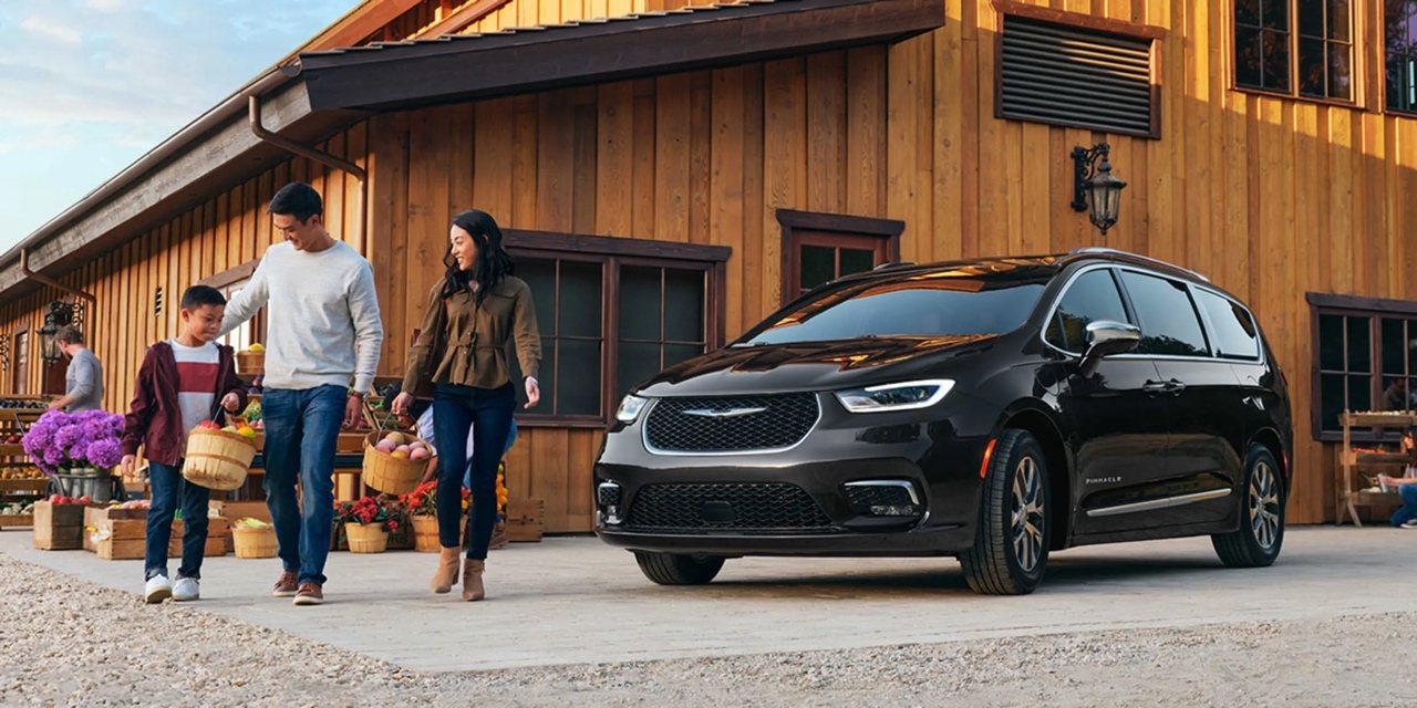 Chrysler Pacifica hybride rechargeable 2026 noire stationnée devant une maison en bois, famille avec enfants transportant des paniers de fruits.