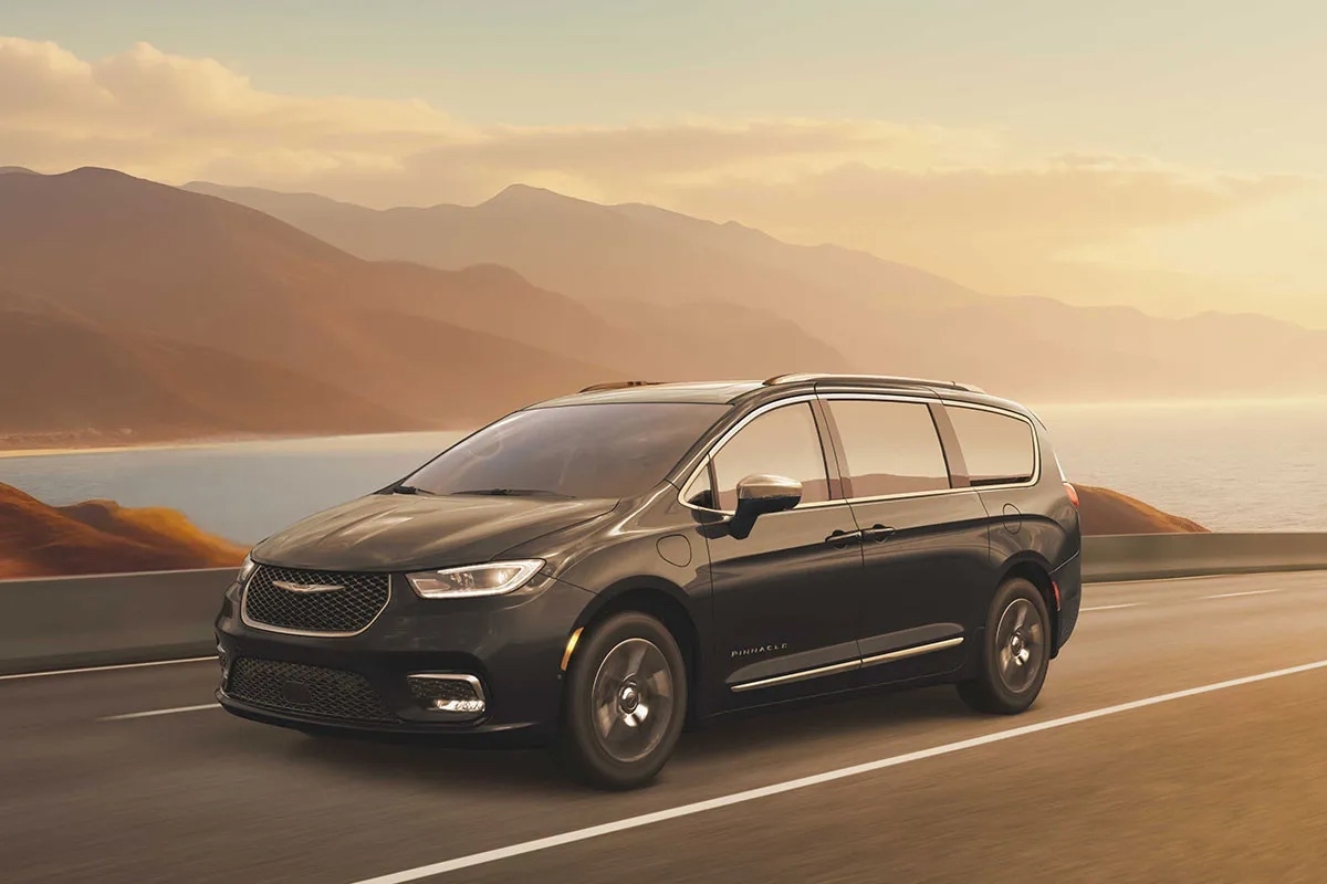 Chrysler Pacifica hybride rechargeable 2026 noire roulant sur une route côtière au coucher du soleil, avec montagnes en arrière-plan.