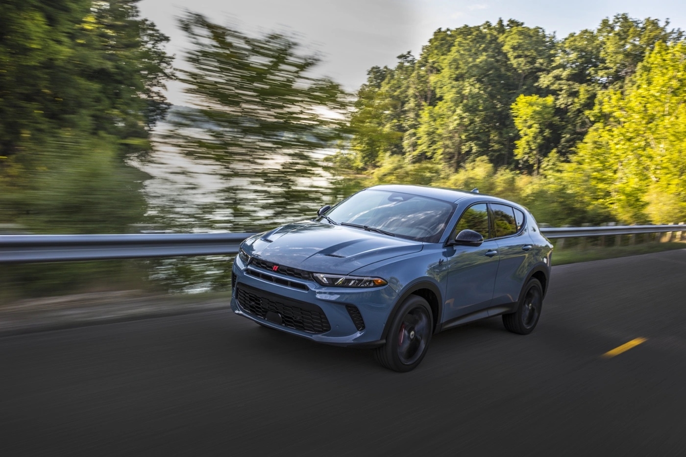 Dodge Hornet 2025 gris en conduite dynamique sur route de campagne bordée d’arbres.